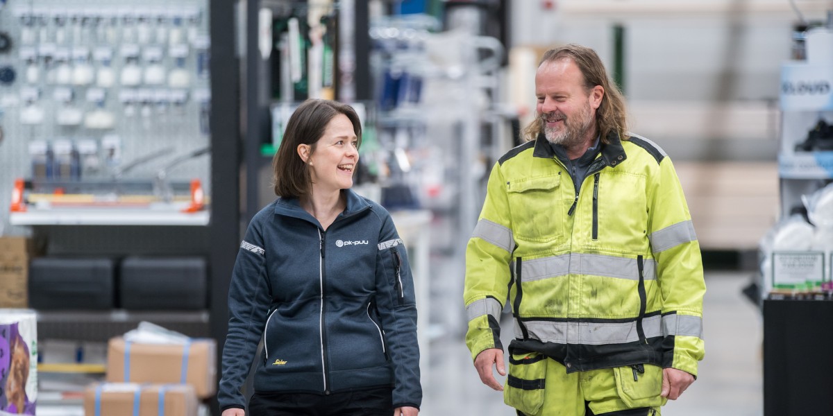 Rautakauppa PK-Puun logotakkiin pukeutunut työntekijä ja huomiovaatteita käyttävä asiakas kävelevät myymälän käytävällä hymyillen ja keskustellen.